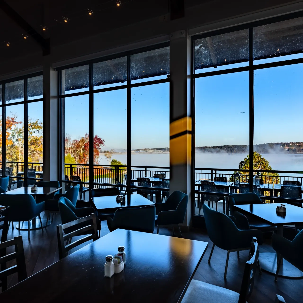 Restaurant interior with large windows overlooking a misty lake and autumn trees.