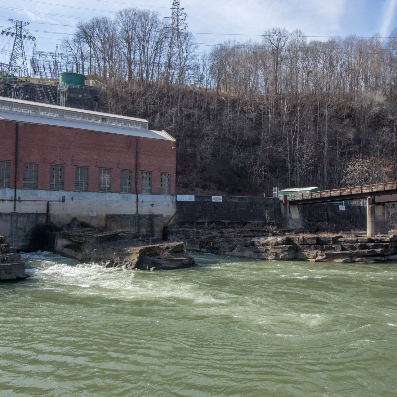 A red brick hydroelectric plant beside a flowing river, bridge to the right, rocky shore, leafless trees, and power lines above, creating a serene, industrial scene.