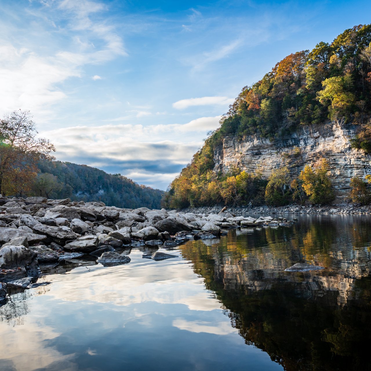 A tranquil river reflects the blue sky and surrounding cliffs, with scattered rocks lining the shore.
