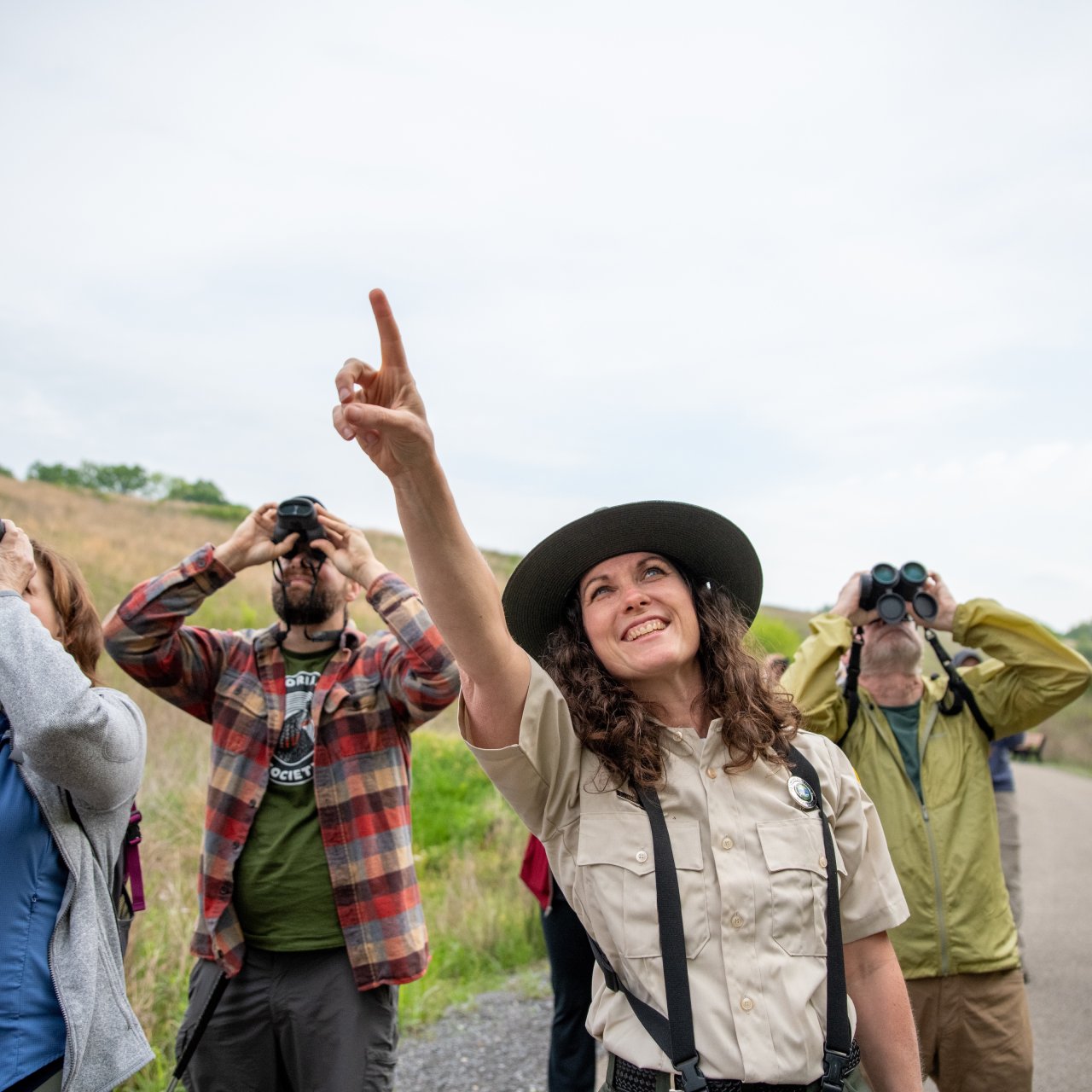 A park ranger in a uniform and wide-brim hat points upward, leading a group of birdwatchers with binoculars on a scenic, overcast nature trail.