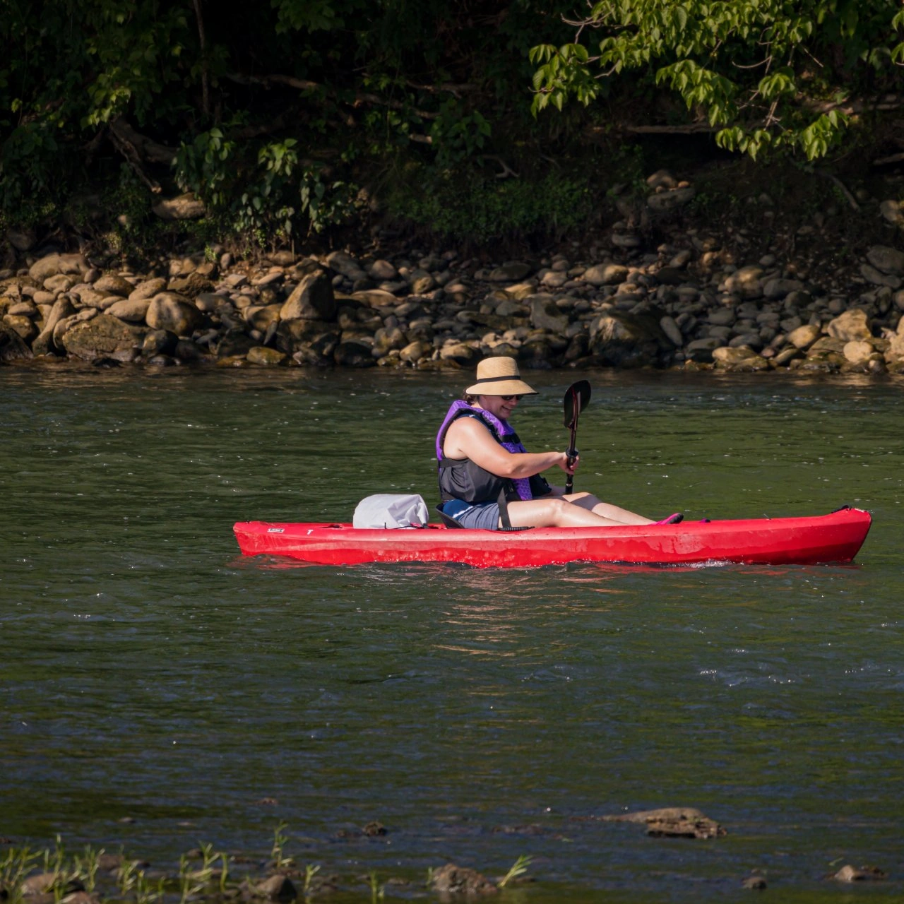 Person kayaking in a red kayak on a calm river, wearing a sun hat and life jacket. The background shows green foliage and a rocky shore, evoking tranquility.