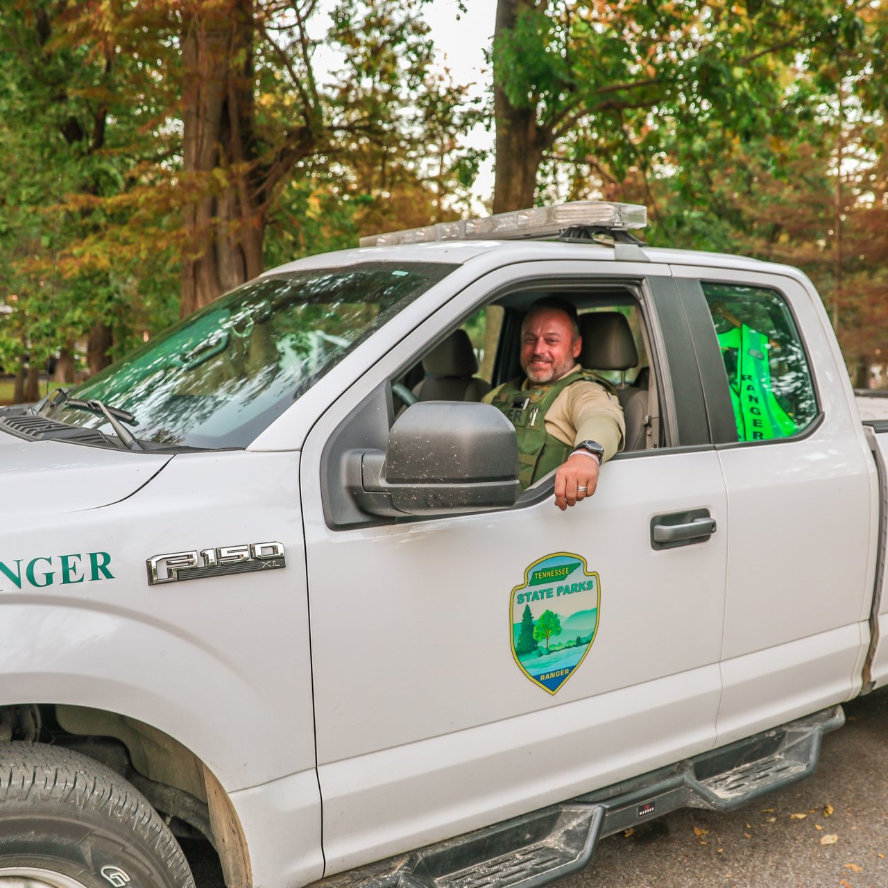 Park ranger in uniform sits smiling in a white truck marked "Park Ranger." The background features lush green trees, creating a serene forest setting.