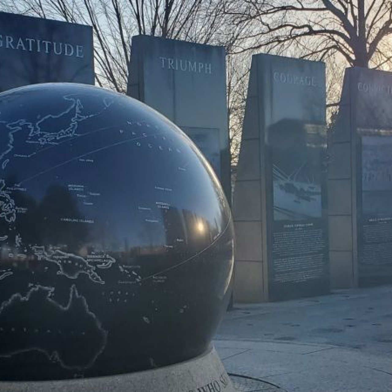 The black granite globe of the World War II Memorial sits in the foreground, with engraved stone panels labeled “Gratitude,” “Triumph,” “Courage,” and more lit by early morning sunlight.