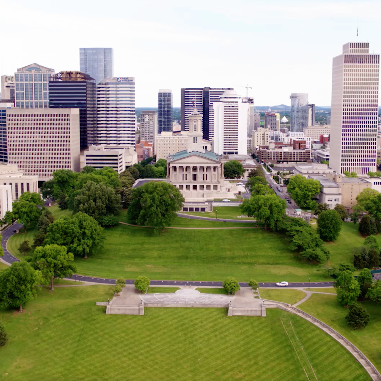 Aerial view of a cityscape featuring a prominent neoclassical building, surrounded by lush green lawns and trees. Tall skyscrapers in the background under a clear sky convey a lively urban atmosphere.