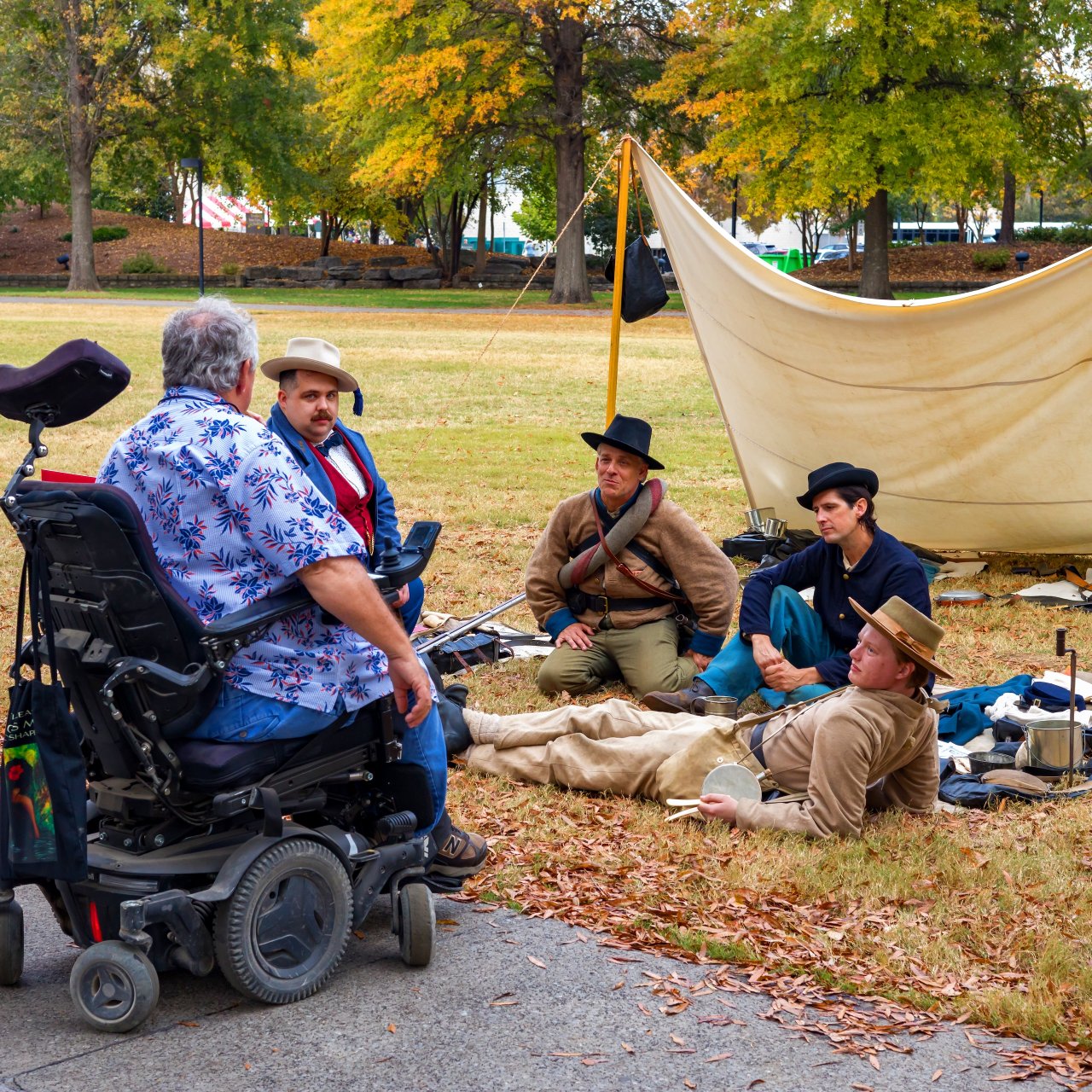A person in a wheelchair talks to four people in historical attire by a canvas tent on grass. The scene is relaxed, with autumn trees in the background.