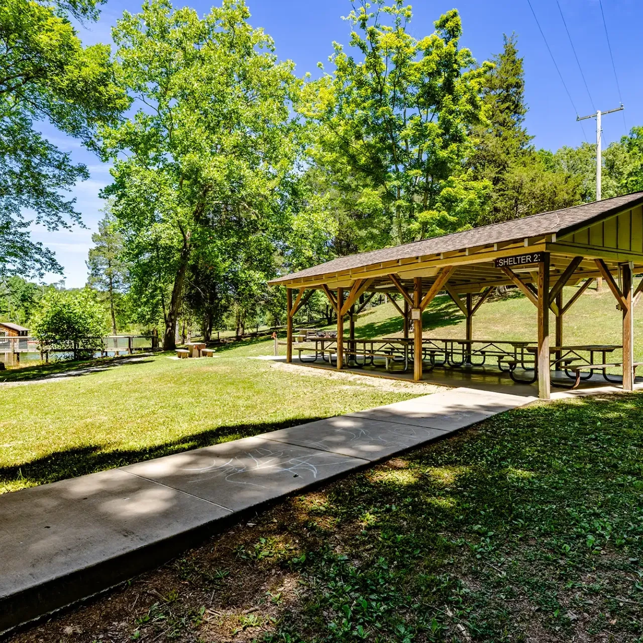 A sunny park scene with a wooden picnic shelter labeled "Shelter 2," surrounded by lush green trees and grass. A concrete path leads to the shelter.