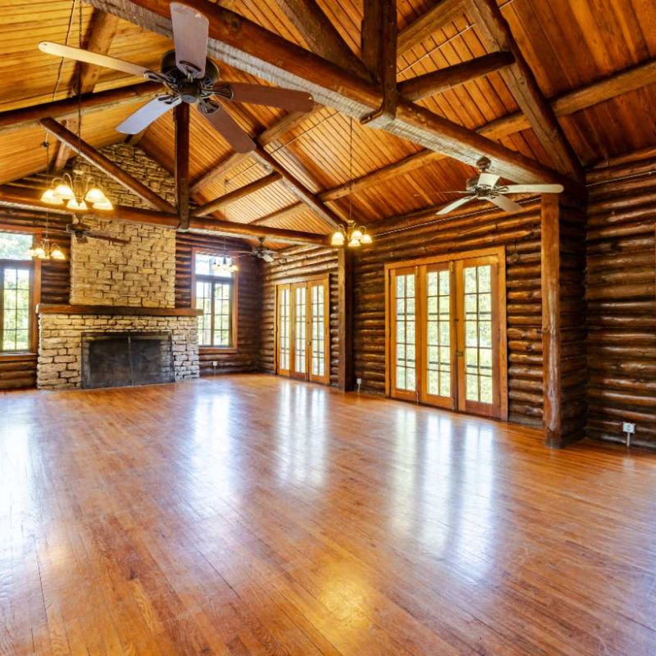 Spacious log cabin interior with wooden beams, stone fireplace, and polished wooden floors. Bright natural light through multiple French doors. Cozy, rustic ambiance.