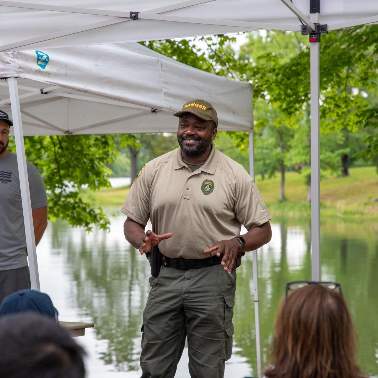 A park ranger speaks animatedly under a canopy near a pond, surrounded by green trees. Attendees listen attentively, creating a relaxed outdoor setting.