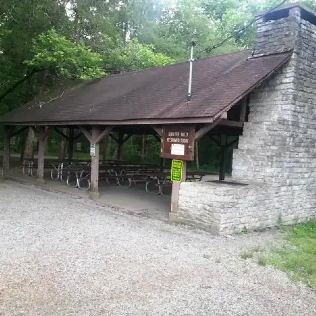 Park pavilion with picnic tables and stone fireplace, surrounded by trees.