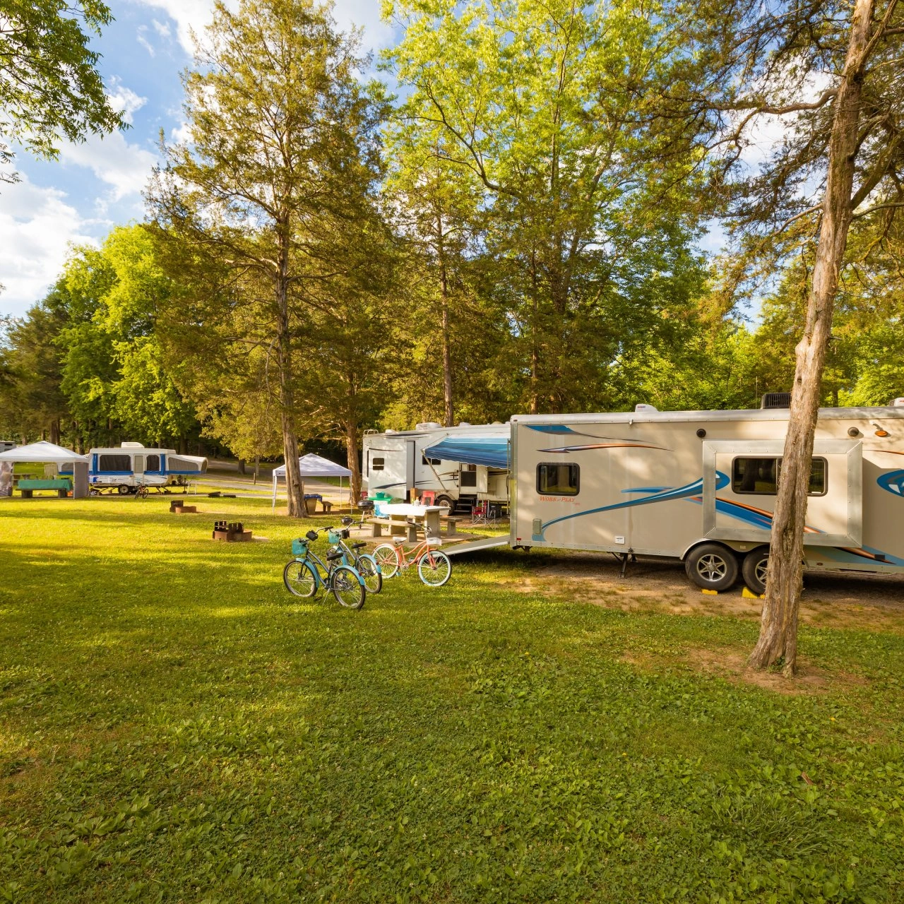 Large RV with bikes in a sunny campsite surrounded by trees.