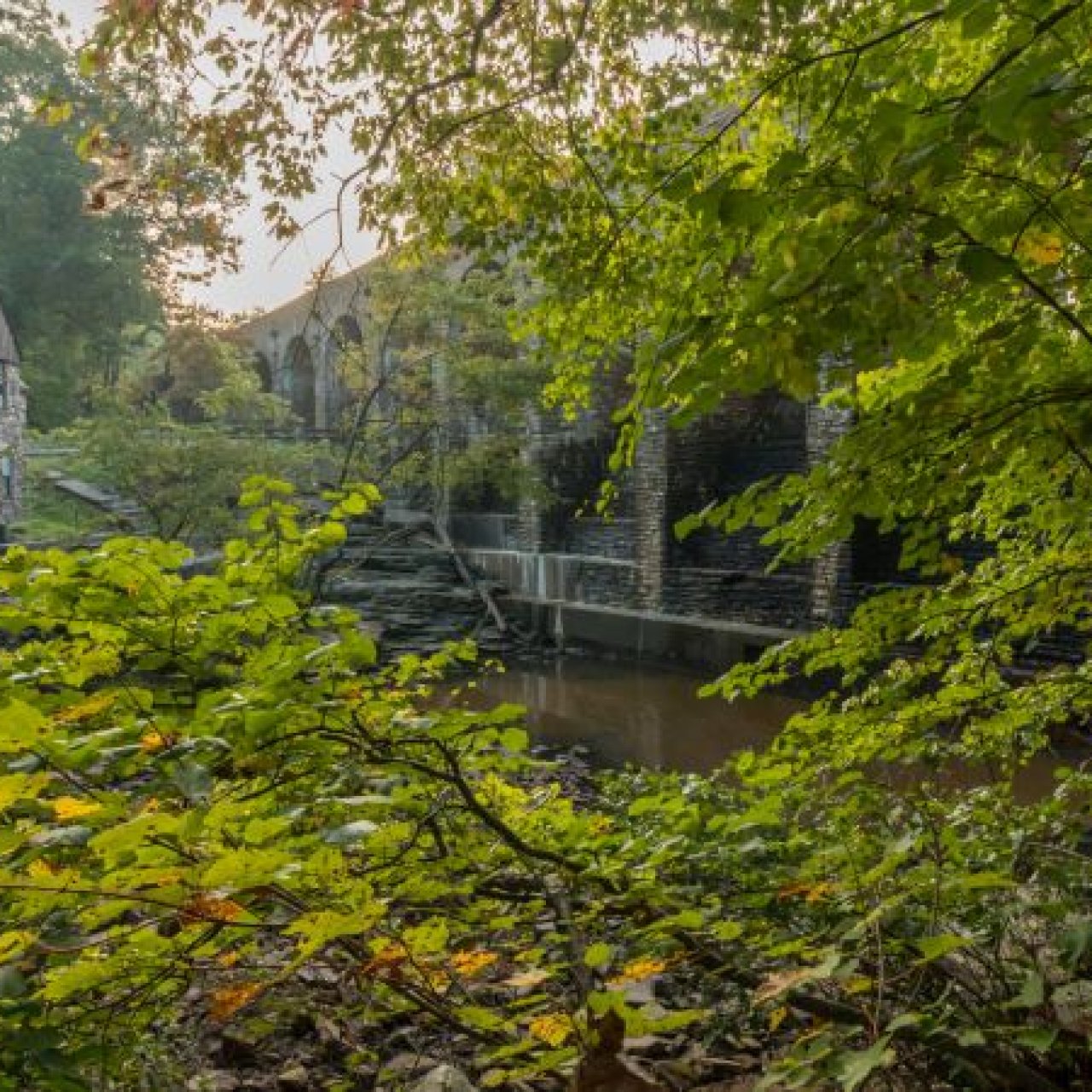 A rustic stone mill building and arched bridge are partially hidden by lush green leaves, with a small waterfall spilling into the stream below.