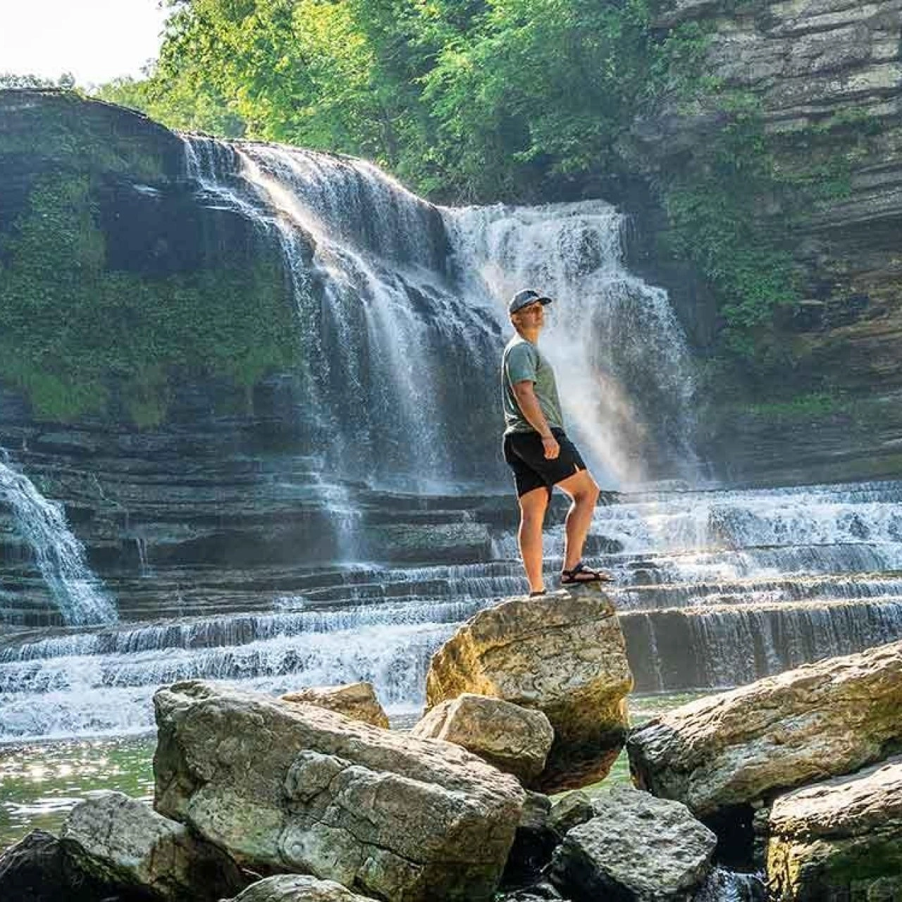 Man standing on rocks by a cascading waterfall, surrounded by lush greenery.