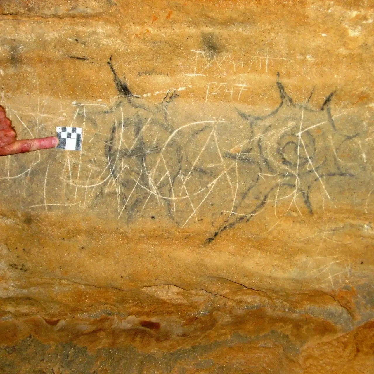 Rock wall with etched patterns, a hand pointing at a small checkerboard for scale.