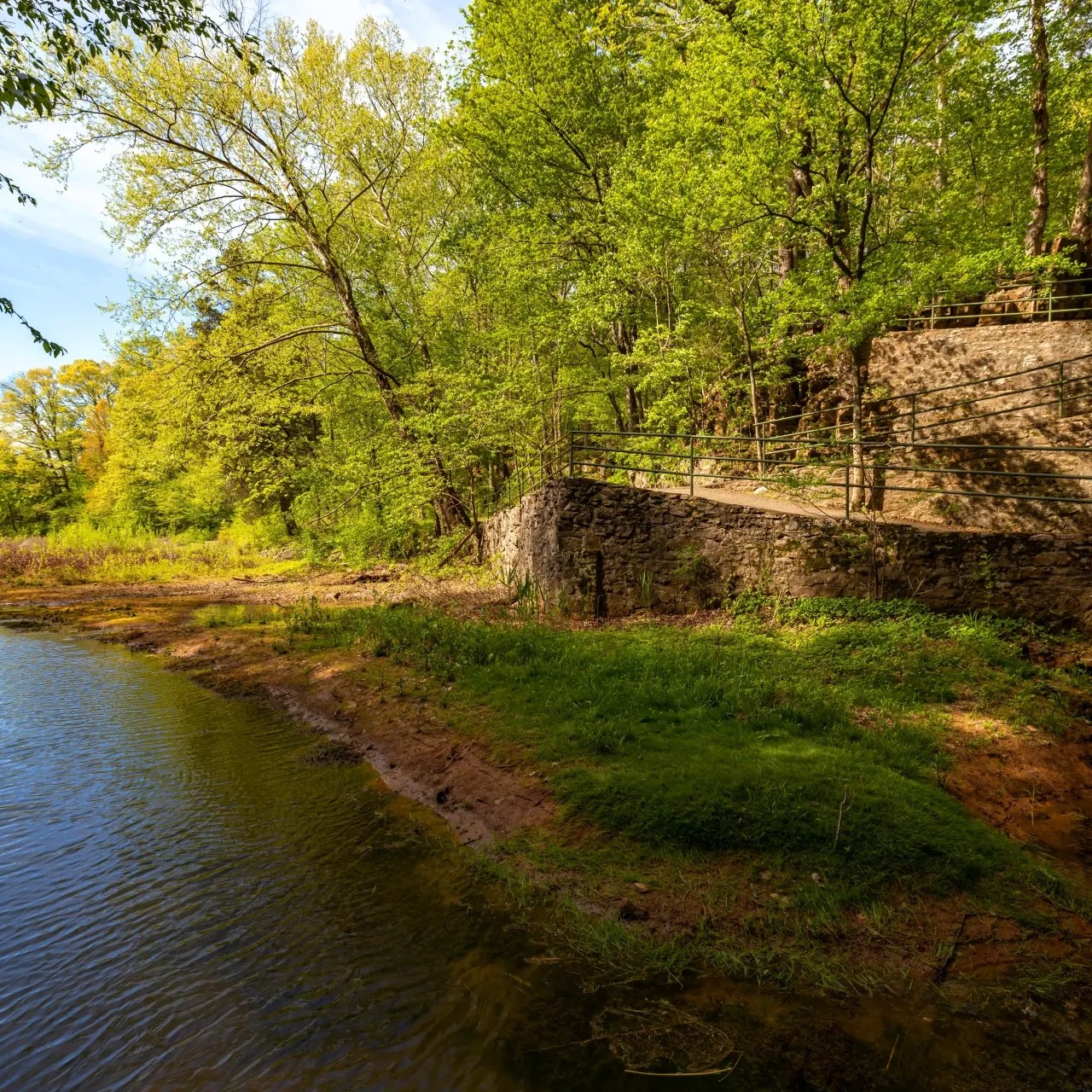 River beside a stone wall and lush green trees under a clear blue sky.