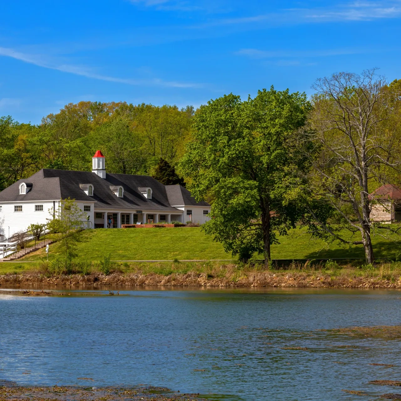White house with red roof by a pond and trees, under a clear blue sky.