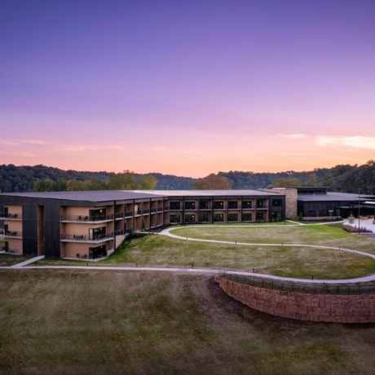 Modern lakeside hotel at sunset with a curved design, surrounded by lush greenery and woodland. The sky is a gradient of purple and orange hues.