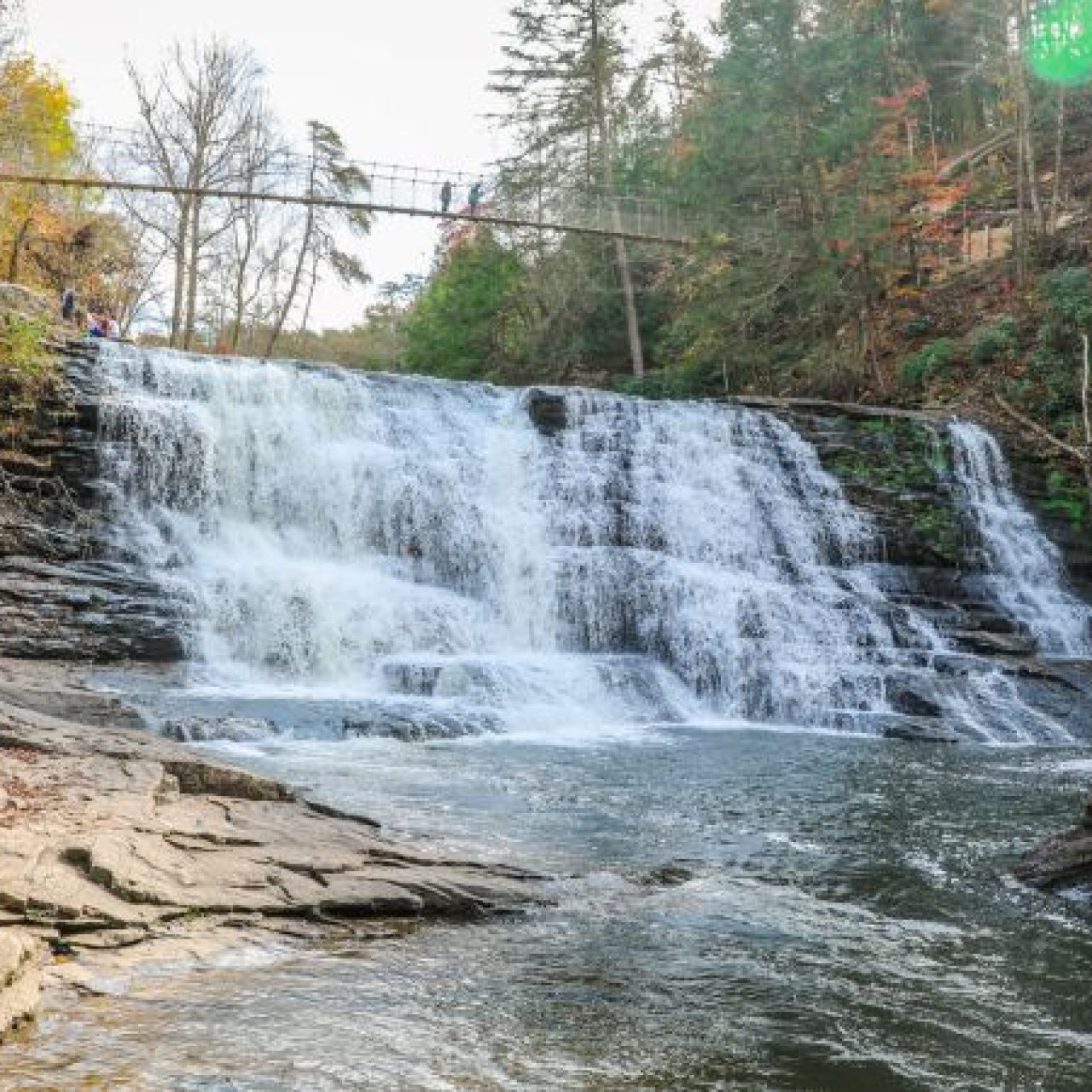 A serene waterfall cascades down layered rocks into a clear pool, framed by autumn trees. A footbridge hangs above, with people enjoying the view.
