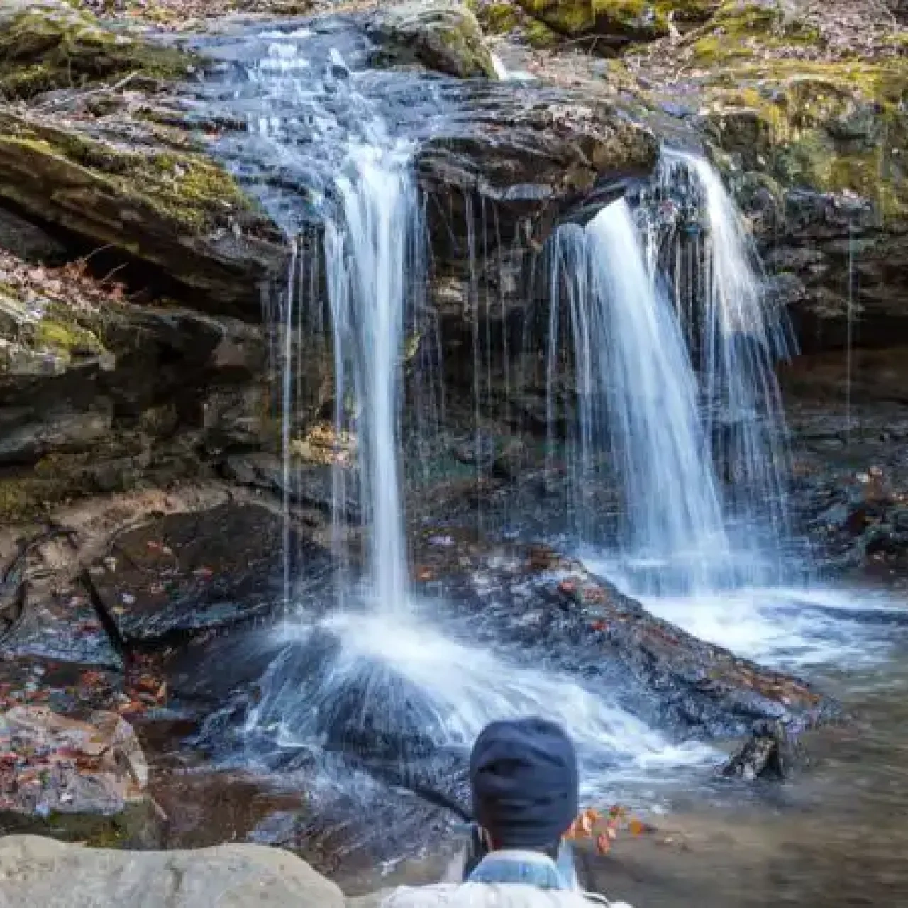 A small waterfall cascades over mossy rocks into a clear pool, surrounded by autumn leaves. A person in a beanie admires the tranquil scene.