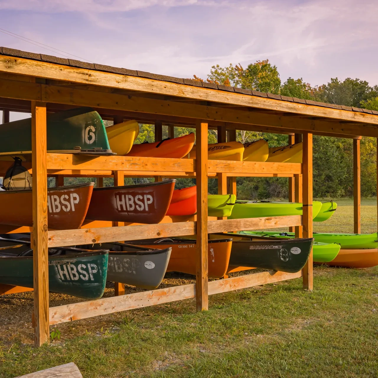 Wooden rack holding colorful kayaks, including green and orange ones, under a roof, set against a backdrop of grass and trees at sunset. Calm, outdoor scene.