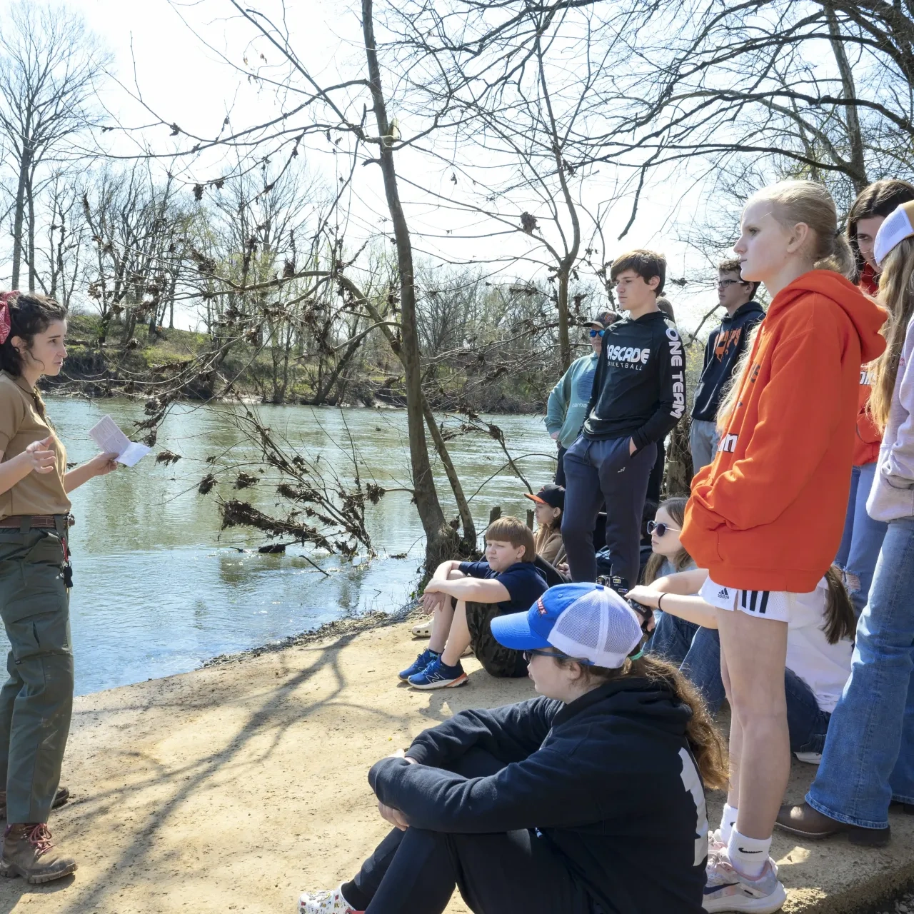 A female park ranger speaks to a seated group of children and teens beside a calm riverbank on a sunny day.