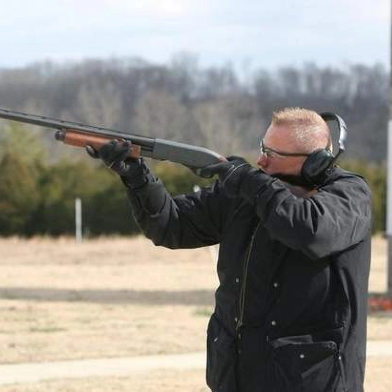 Man wearing ear protection aiming a shotgun at a trap shooting range.