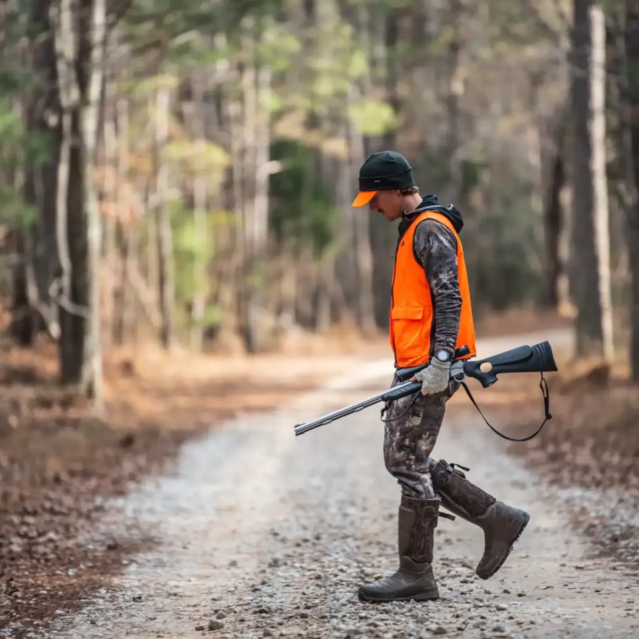 Hunter in orange vest walking on a forest path, carrying a rifle.