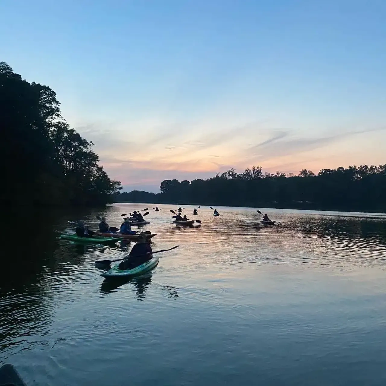 Kayakers on a calm river at sunset, surrounded by silhouettes of trees.