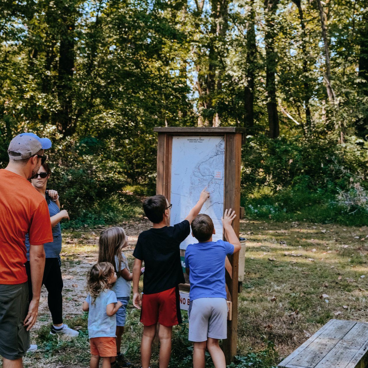 A group of children and two adults gather around a wooden trail map in a forest.