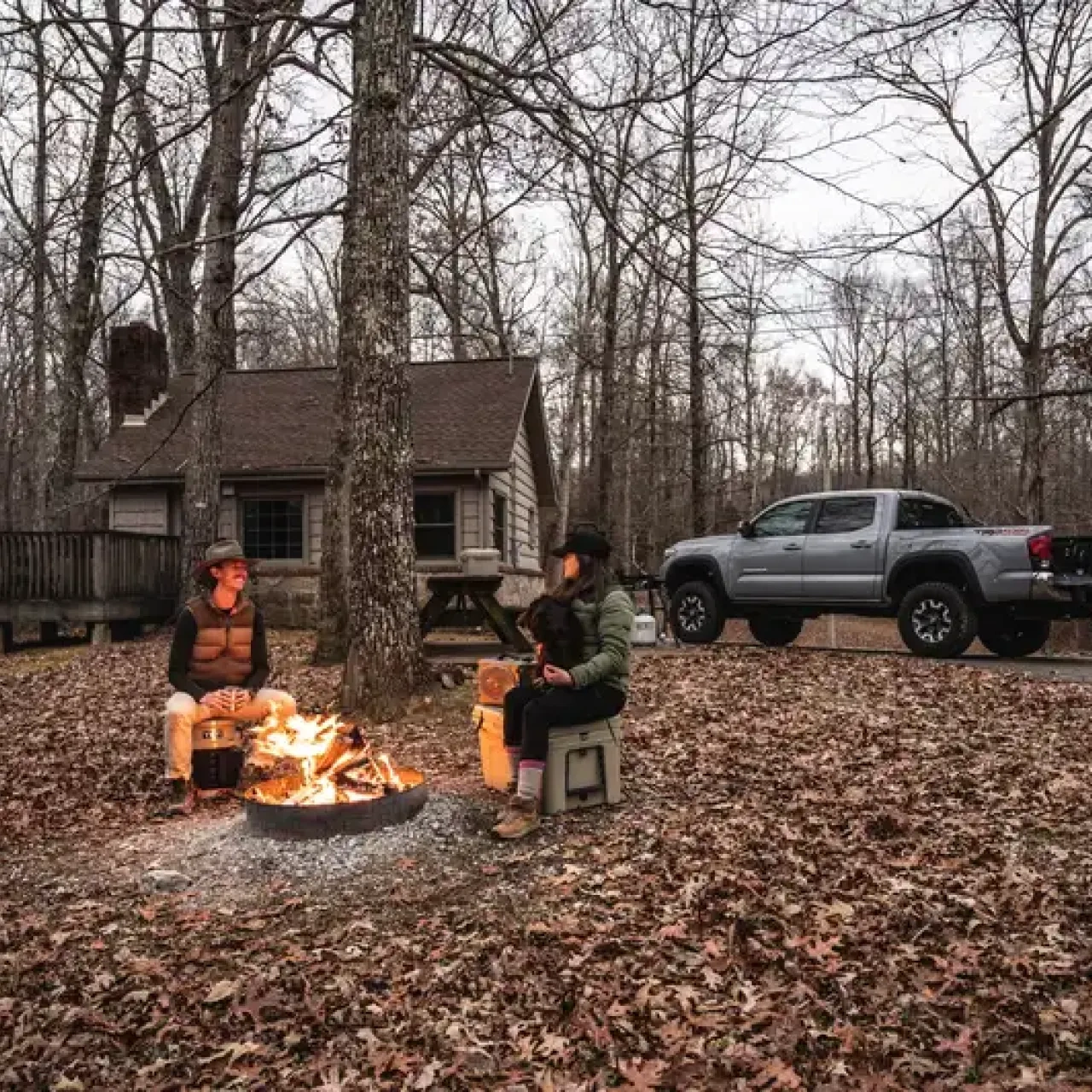 Two people sitting by a campfire near a cabin in the woods, truck parked nearby.