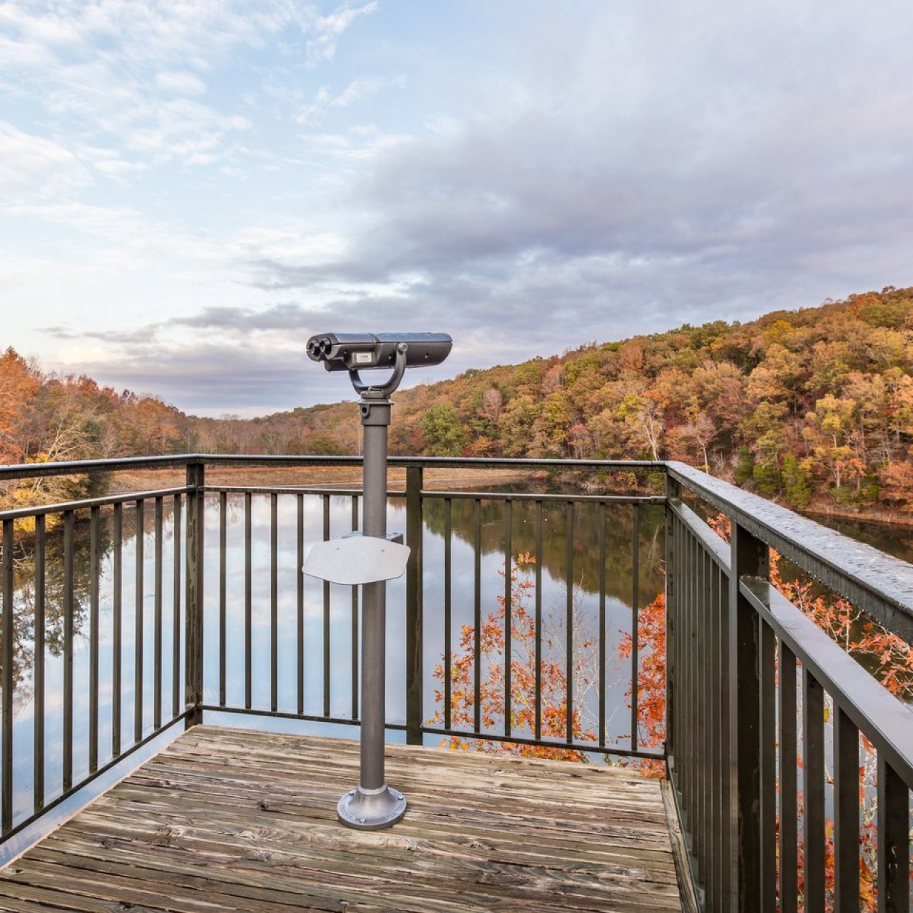 Viewing platform overlooking a calm lake surrounded by colorful autumn trees. A binocular telescope is mounted on the wooden deck