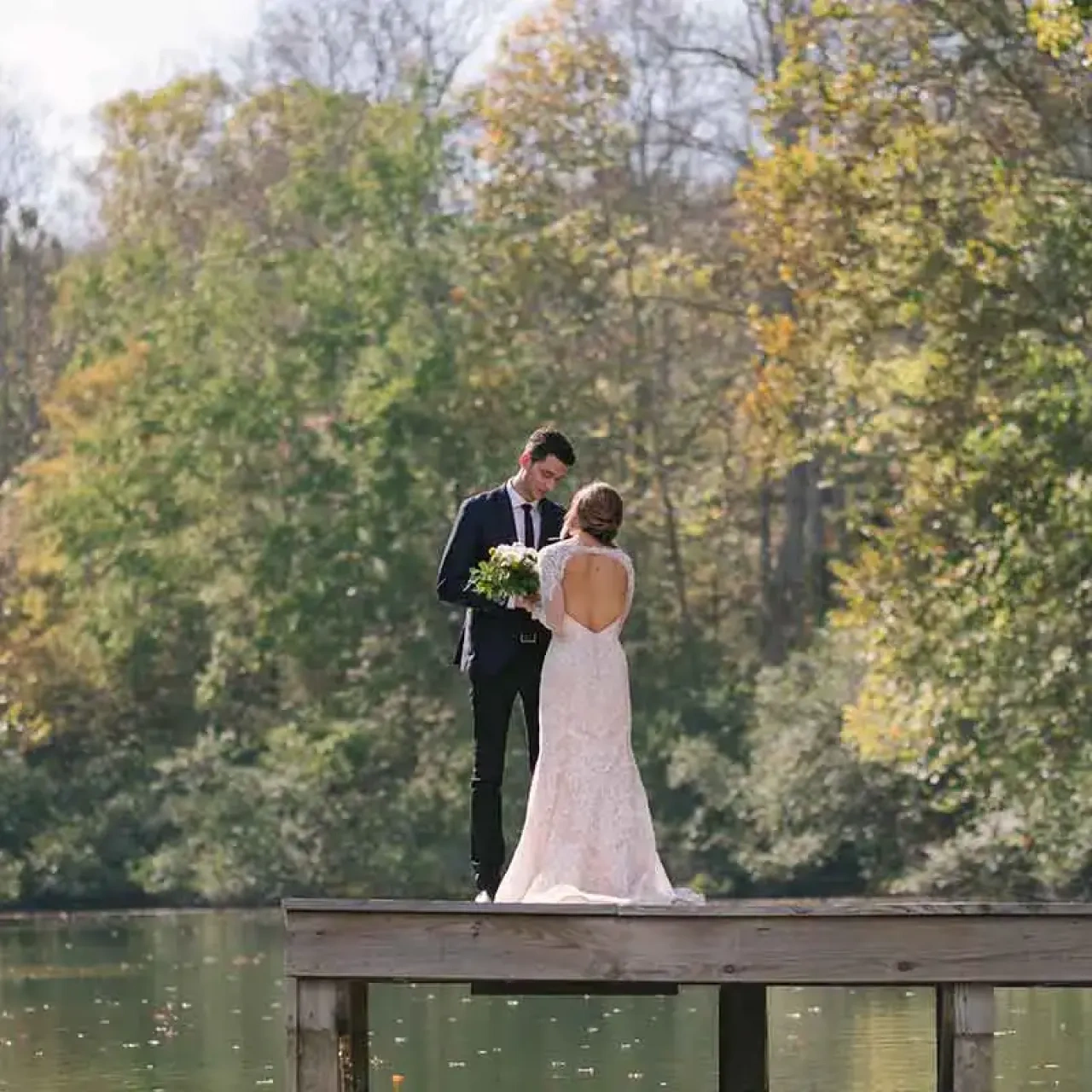 Bride and groom stand on a wooden dock, surrounded by trees and a calm lake.