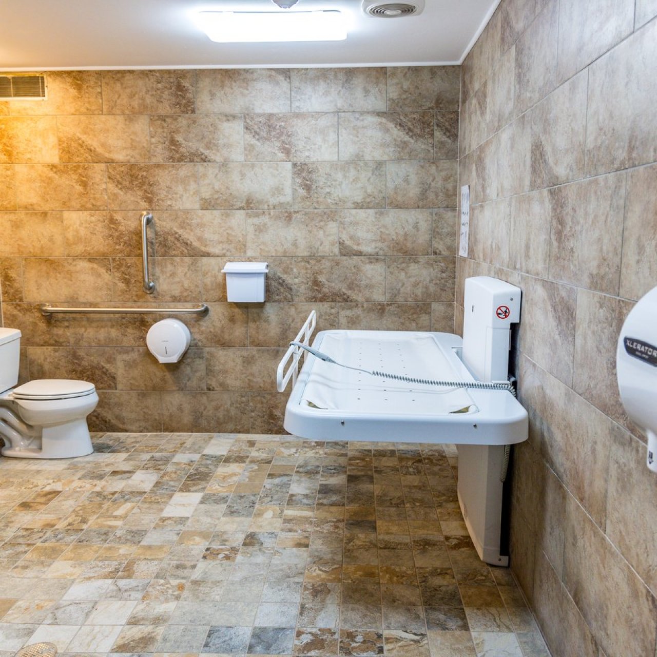 Spacious accessible restroom featuring a toilet, sink, handrails, a wall-mounted changing table, and bright overhead lighting.