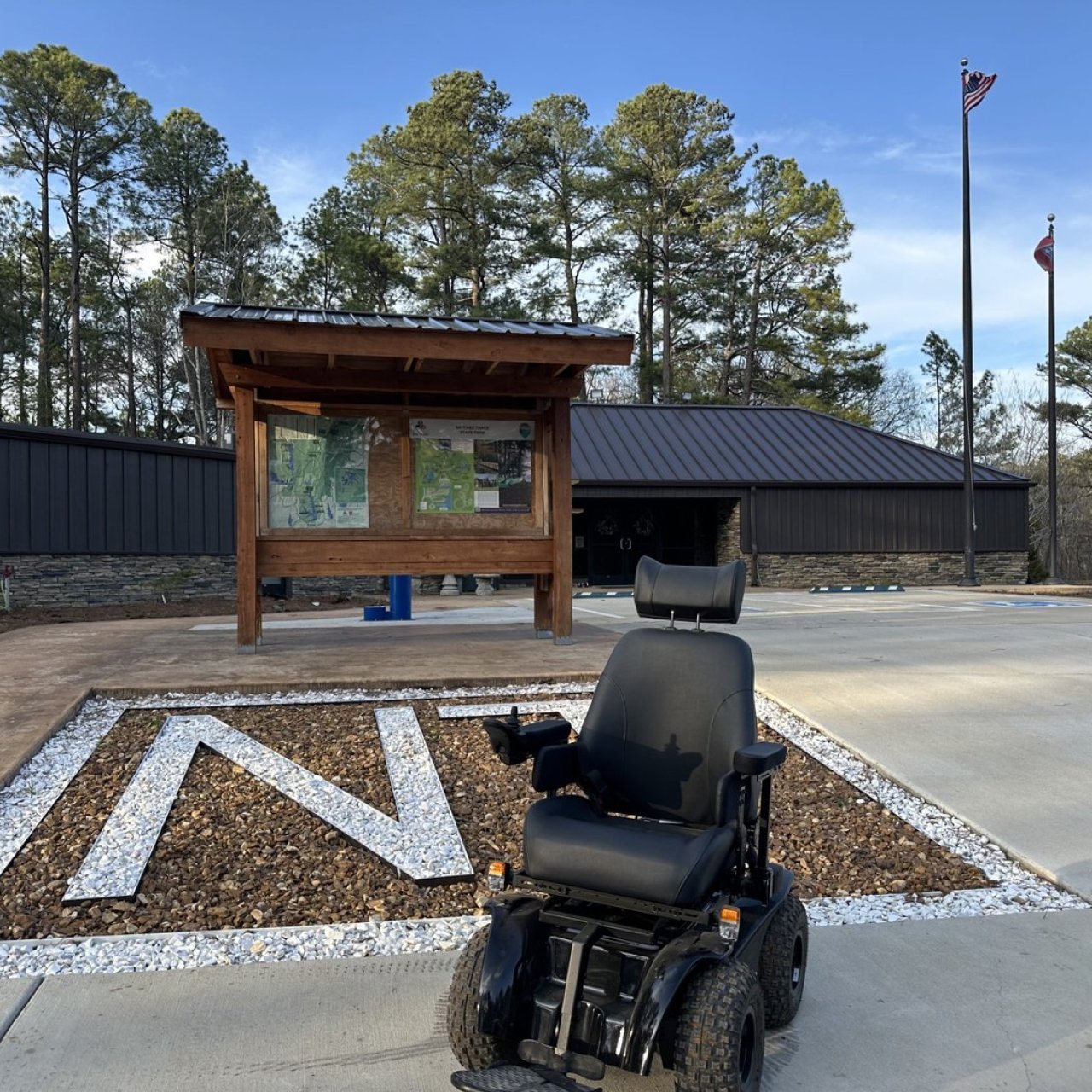 A black electric wheelchair is parked near a wooden information board outside a building, with a paved area featuring a large "N". 