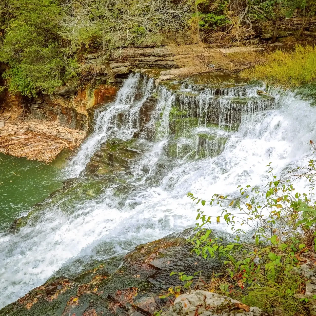 Waterfall cascading over rocks, surrounded by greenery and trees.