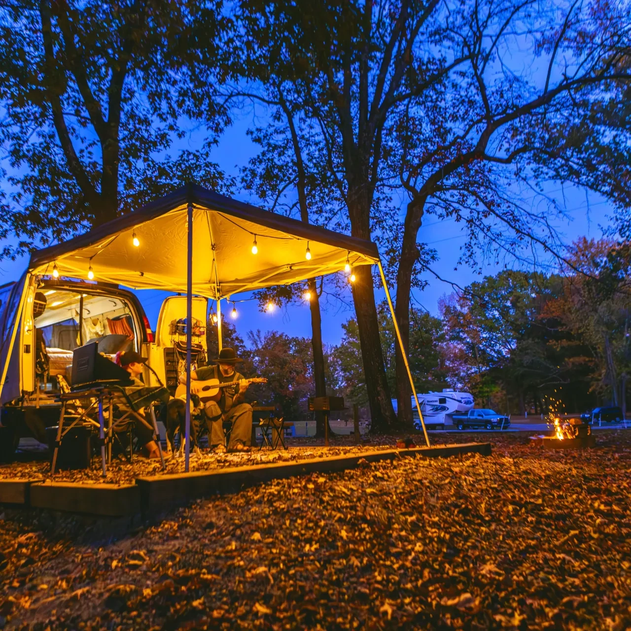 Van campsite at dusk, warm lights, people playing guitar, surrounded by autumn trees.
