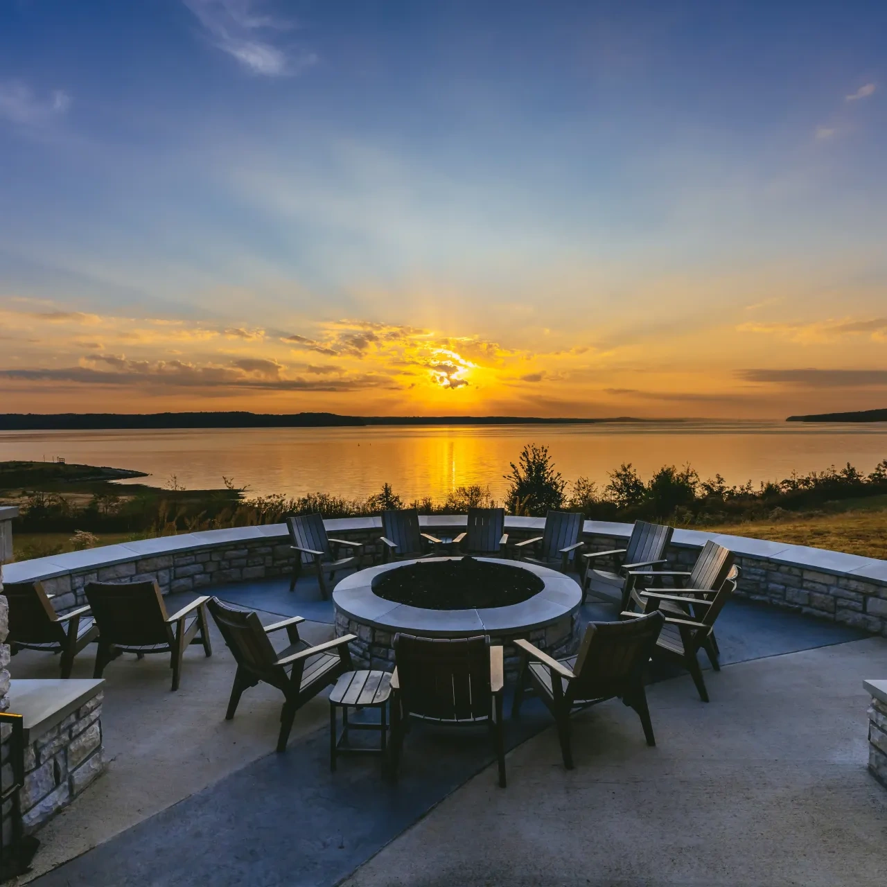 Sunset over lake with stone fire pit and chairs in foreground.