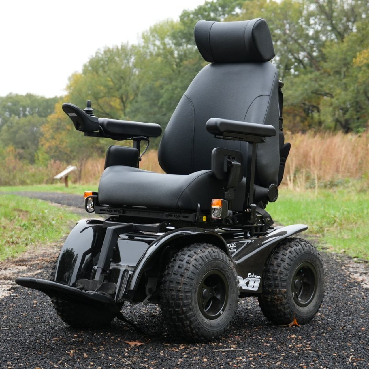 A rugged black power wheelchair sits on a winding gravel path in a lush park with tall grass and trees, conveying adventure and independence.