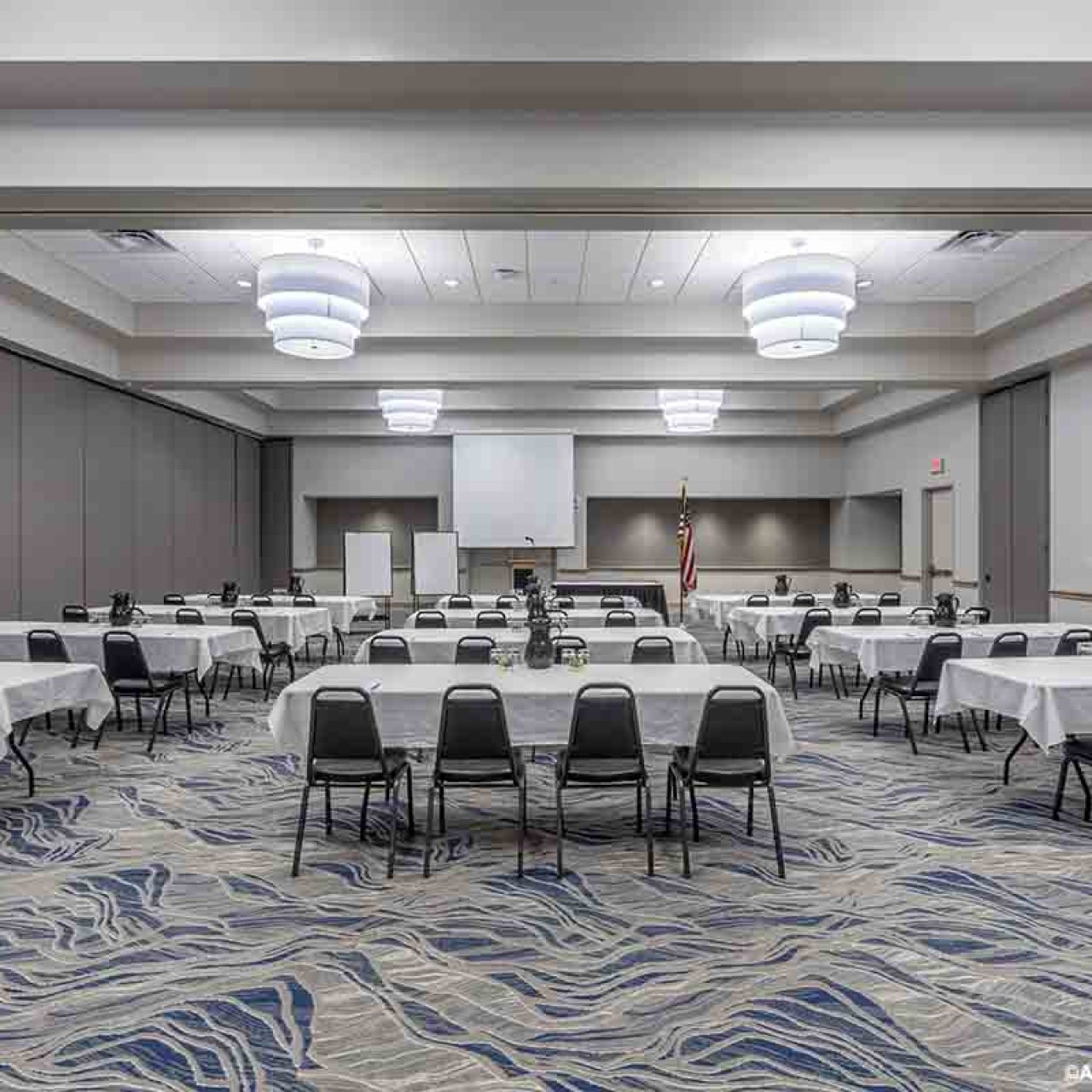 Large room with tables with white cloth set up with chairs, the room has a grey carpet with blue patterns and overhead lighting