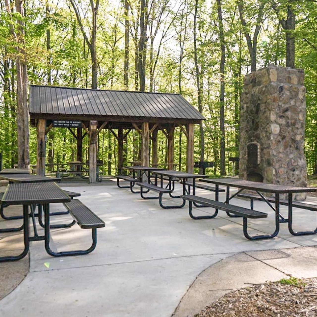 small open concreted area with six metal picnic benches, a pavilion, and a stone furnace