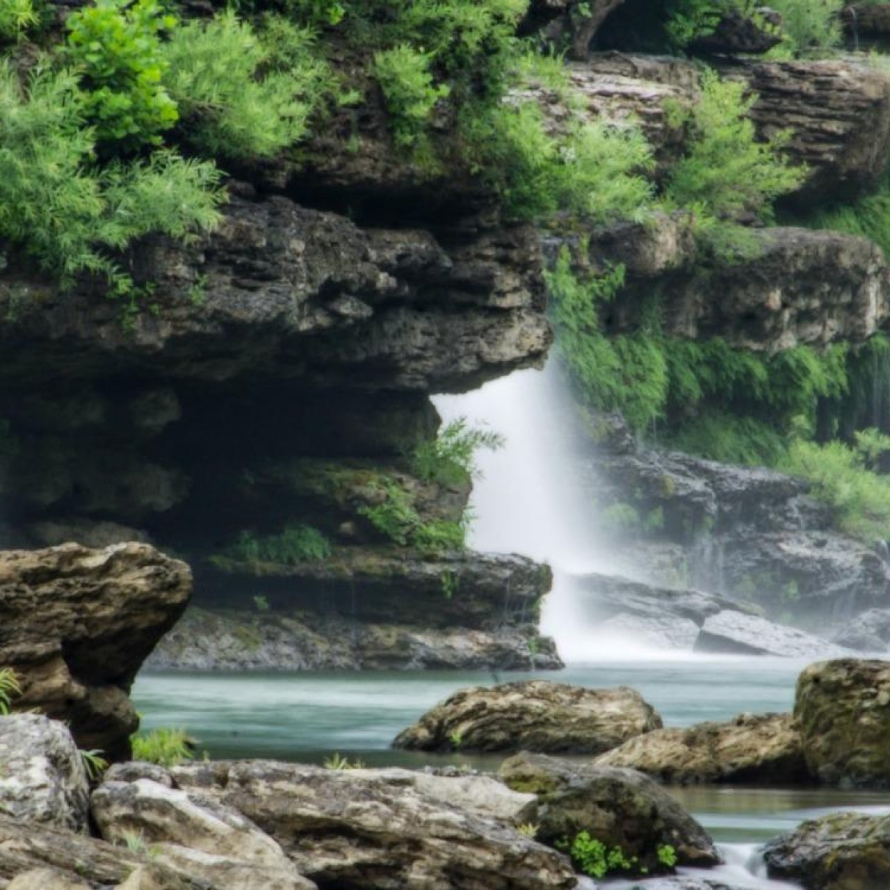 A serene waterfall cascades over mossy rocks into a calm pool, surrounded by lush green foliage. The scene conveys tranquility and natural beauty.