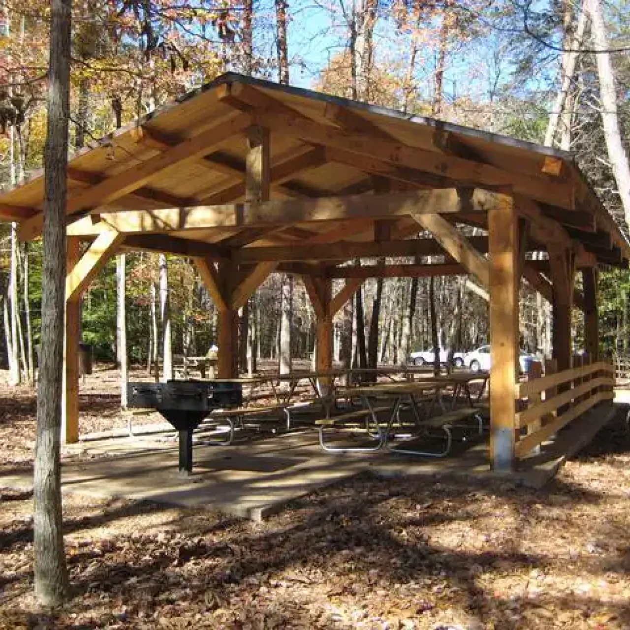 Wooden picnic pavilion in an autumn forest setting. Several picnic tables are underneath, with a grill nearby. Sunlight filters through colorful leaves.
