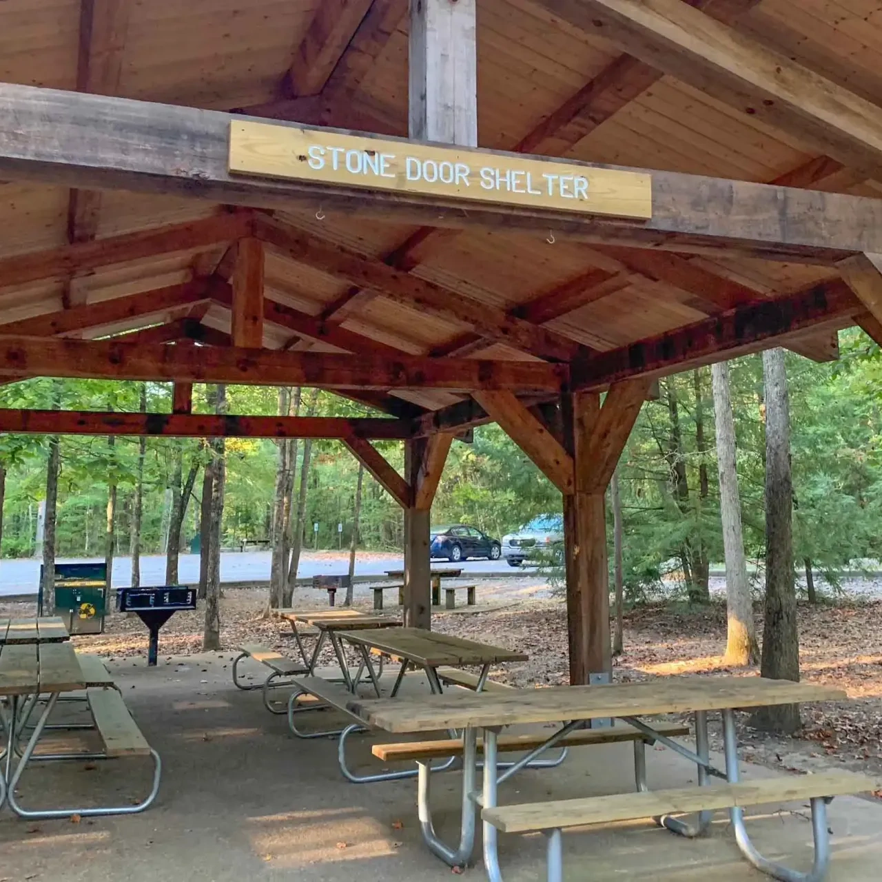 Wooden pavilion labeled "Stone Door Shelter" with picnic tables underneath. Surrounded by trees and a parked car in the background on a sunny day.