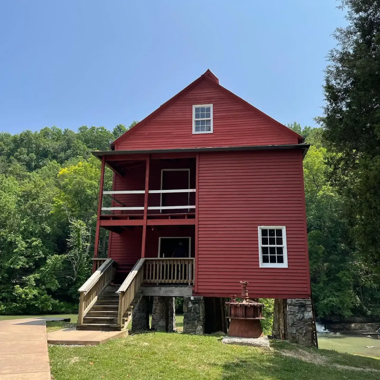 A tall, red barn-like building with a pitched roof stands on stone pillars. It features a front porch with stairs and is surrounded by lush green trees under a clear blue sky.