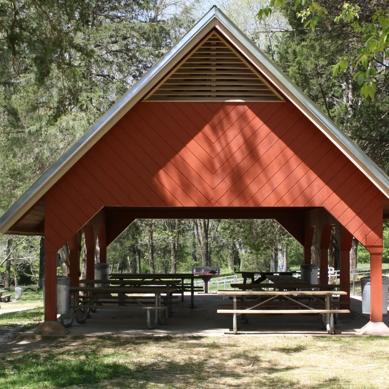 A red picnic shelter with a peaked roof stands in a wooded park. Several picnic tables are underneath, surrounded by tall green trees. Calm and inviting.