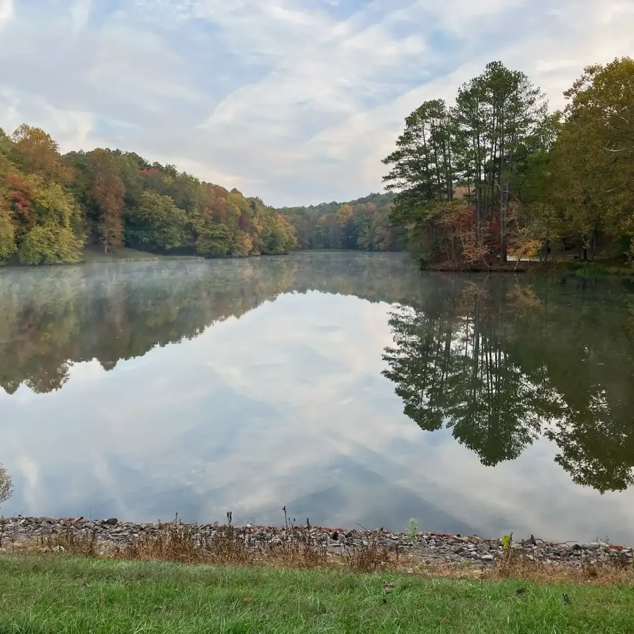 Calm lake with autumn trees reflecting on the water, grassy foreground.