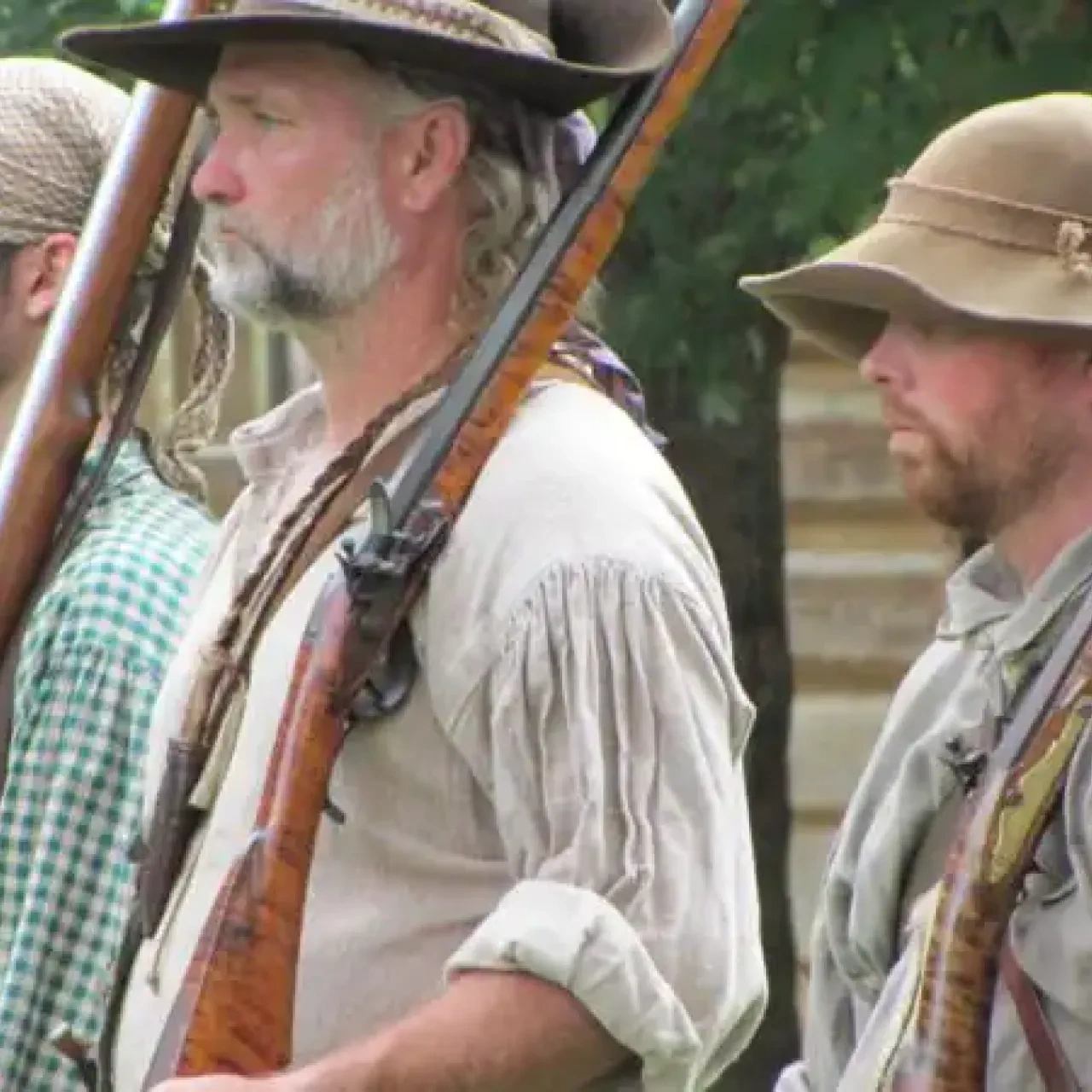 A group of men in historical attire stand in line, holding rifles. They wear hats and period clothing, evoking a rustic, serious atmosphere.