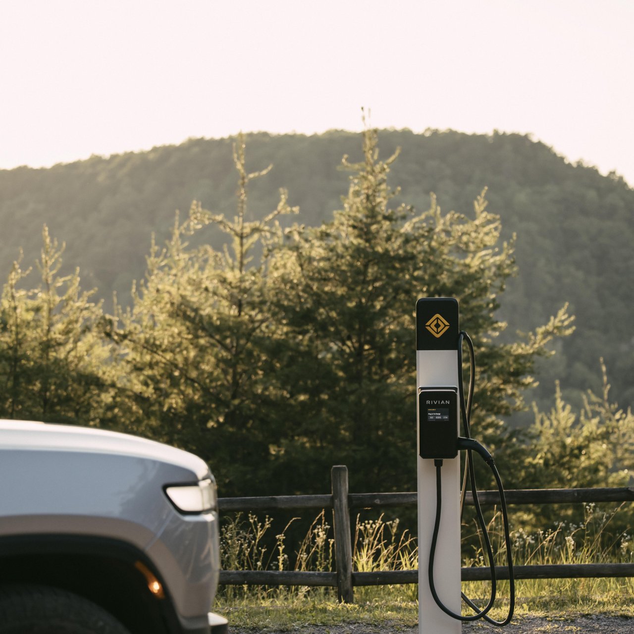 Silver electric vehicle next to a charging station in a scenic landscape with trees and a mountain at sunset, conveying tranquility and eco-friendliness.