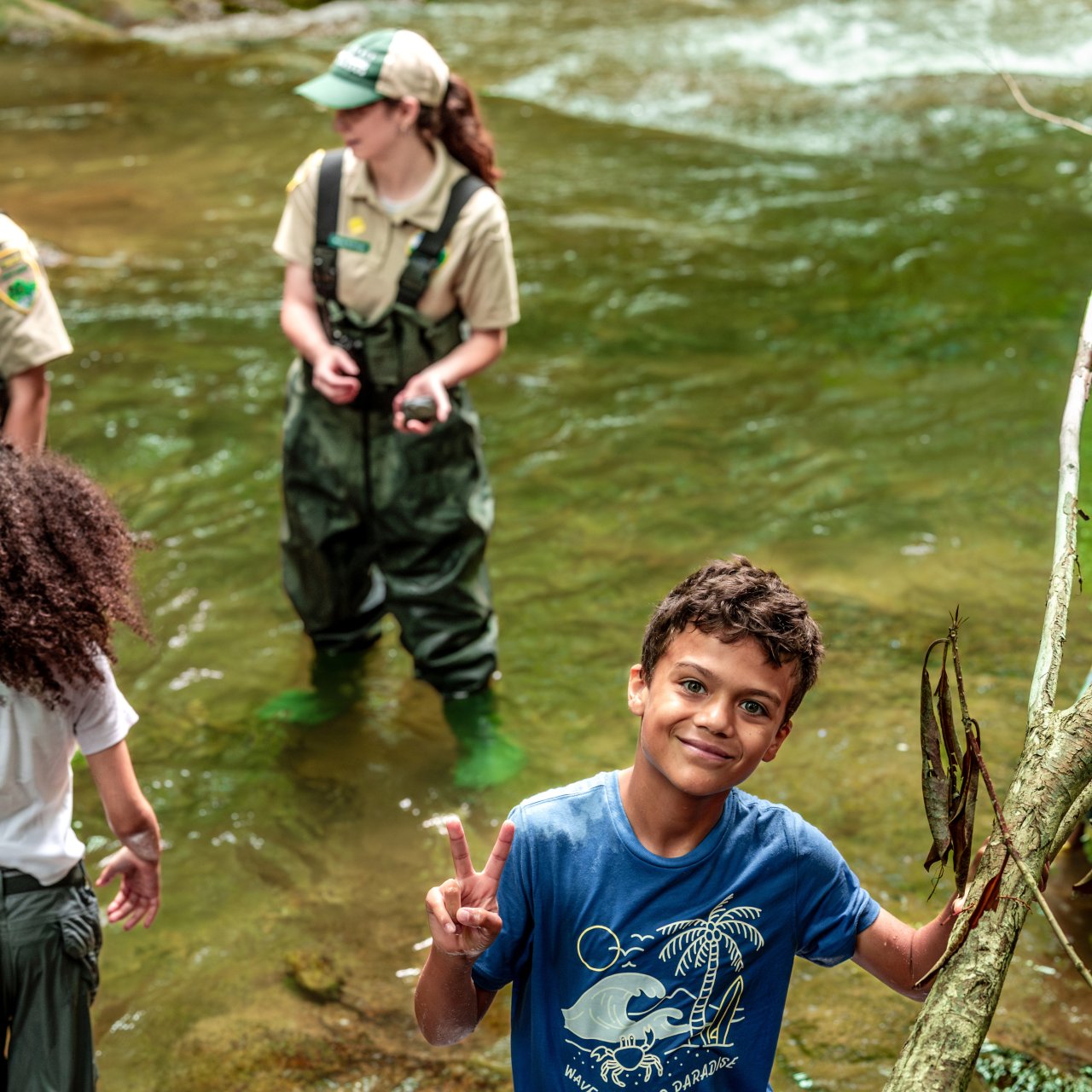 A smiling boy in a blue shirt holds a peace sign beside a stream. A park ranger and another child in shorts wade nearby. The scene is lively and joyful.