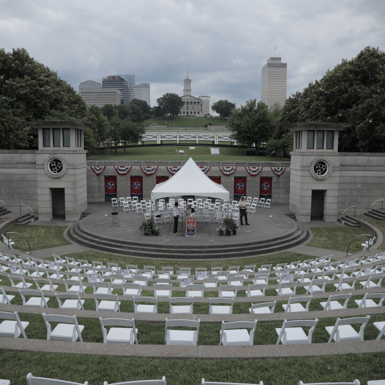 Outdoor amphitheater with white chairs and a stage with a canopy, distant buildings in background.