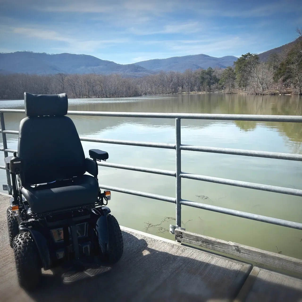 Motorized wheelchair by a lakeside pier, mountains in the background.